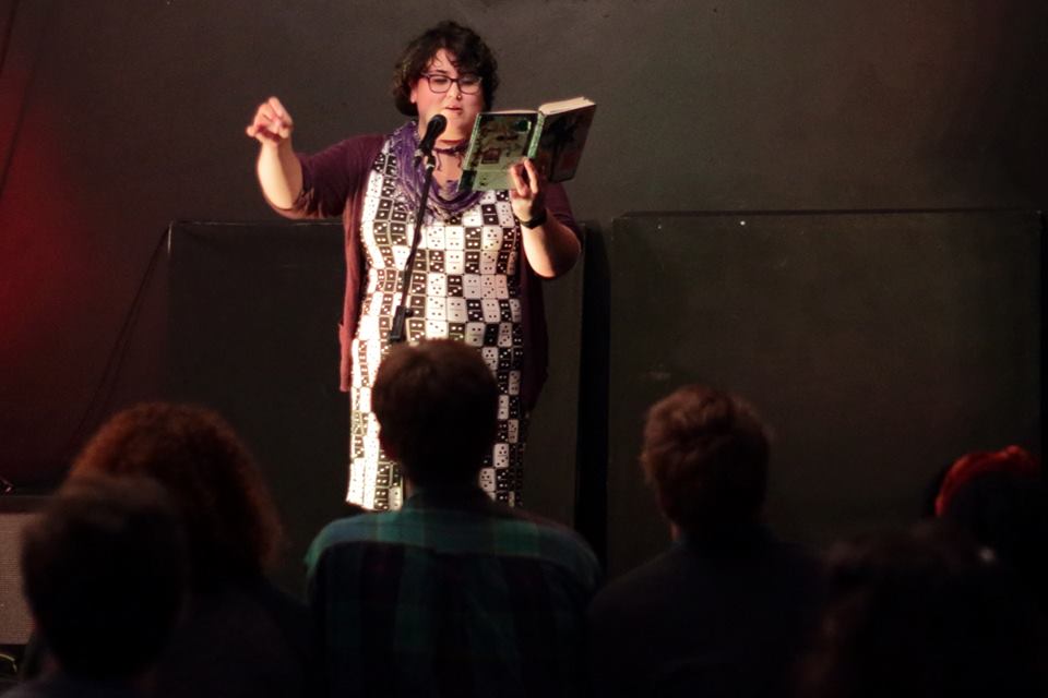 Audience-perspective picture of Cynthia on stage. She is a fat brown latinx femme, reading from her notebook and doing a gesture with her other hand on the microphone. She has short hair and is wearing a domino dress, a purple cardigan and glasses.