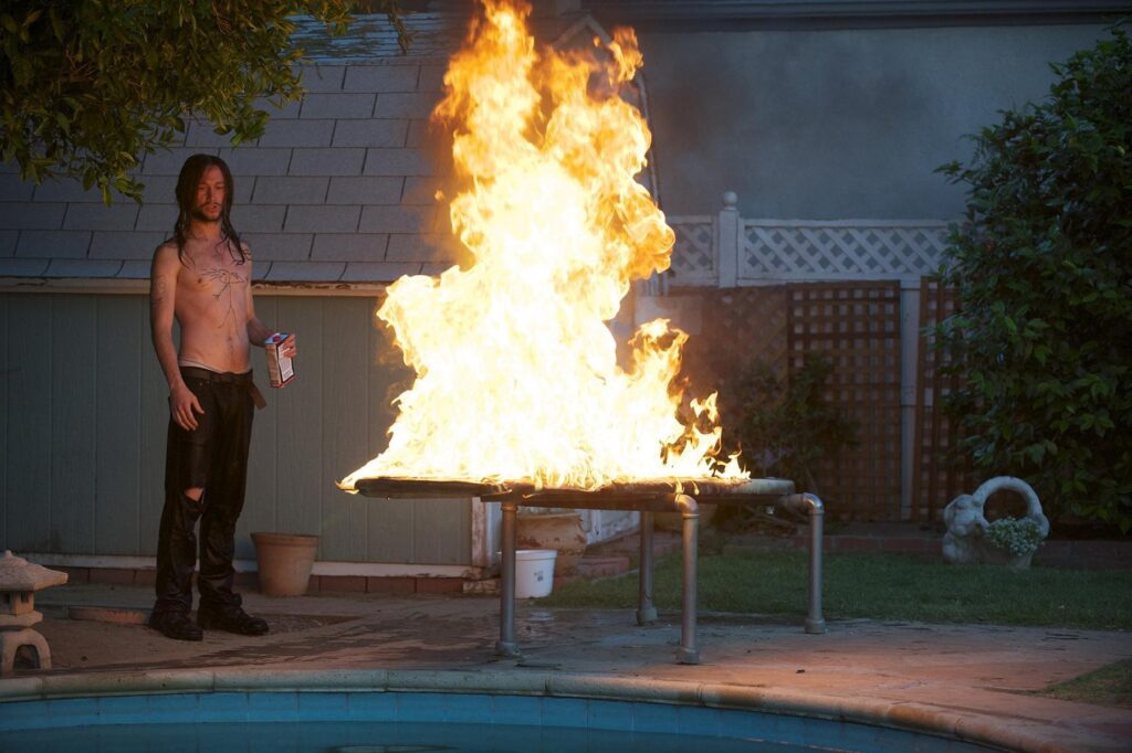 Joseph Gordon-Levitt como Hesher en la película del mismo nombre. Tiene el cabello largo, no trae camisa, y lleva unos pantalones negros rasgados y botas negras. Está en el patio de una casa avejentada con una alberca redonda y sucia. Hay un trampolín oxidado en llamas. Hesher tiene en la mano un bote de material inflamable, como sí él le hubiera prendido fuego al trampolín. Observa la llama despreocupado y tranquilo.