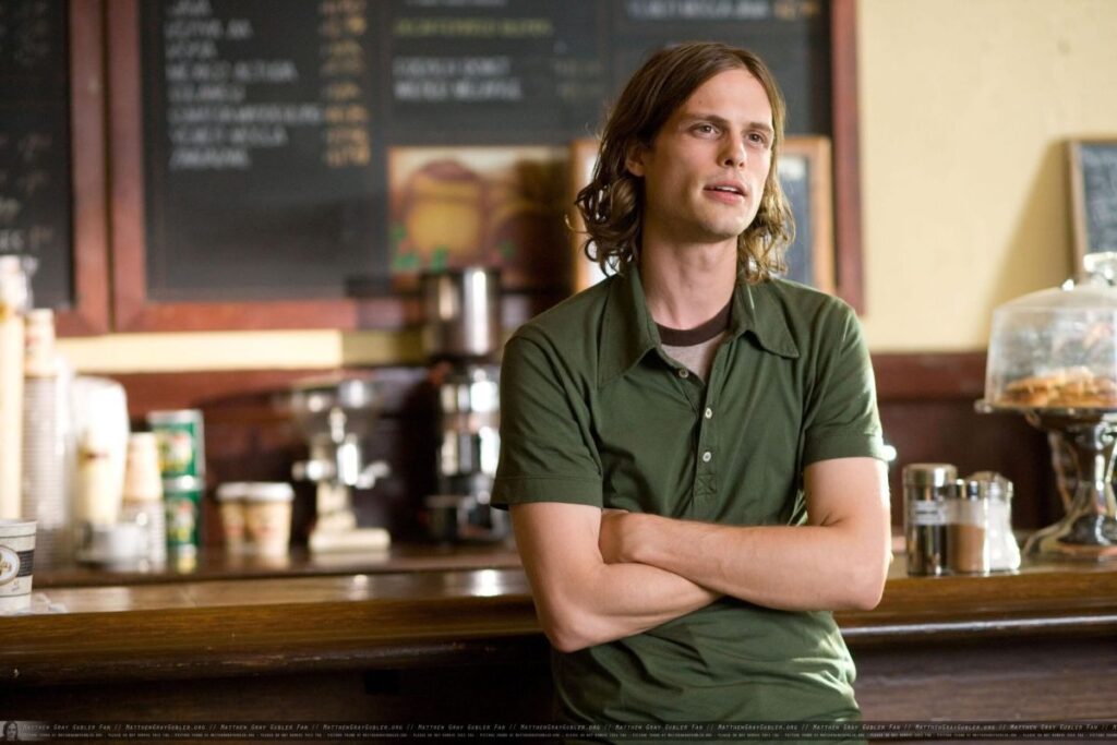 Matthew Gray Gubler como Paul en (500) Days of Summer. Está en una cafetería recargado contra el mostrador, conversando tranquilamente con alguien.